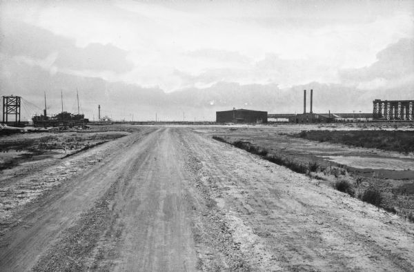 View down a flat, countryside dirt road, and a sulfur factory plant in the distance.