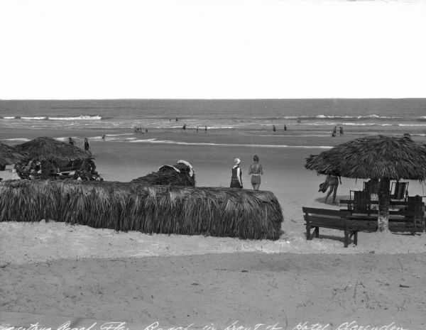 View of the beach in front of the Hotel Clarendon, featuring swimmers, benches, and shade-structures made with palm leaves.