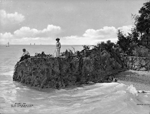 A view of a man fishing from a rock that protrudes into a body of water, with a woman standing behind him, and sail boats are on the horizon.