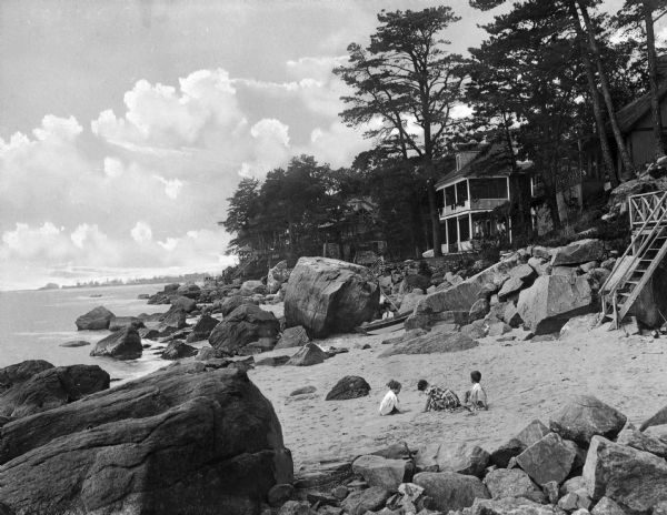 View toward children playing on a beach among large rocks, with beach houses and trees in the background.