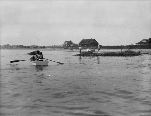 View across water toward the shore. People are rowing a boat in the foreground, and people are standing on a rock near the shore. In the background are houses along the shoreline.