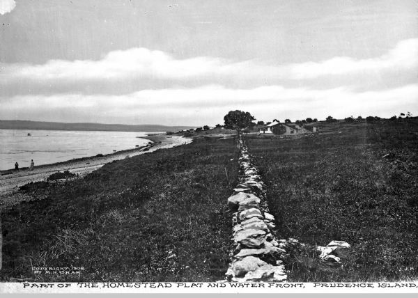 A view of a shoreline. Caption reads: "Part of the homestead plat and waterfront. Prudence Island." Along the left is a rock wall, and a house in the distance. On the left people are walking on the beach.