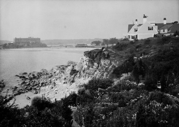 View along top of rocky cliff toward a home on the right overlooking the ocean, with a large building further down the curving shoreline in the distance.