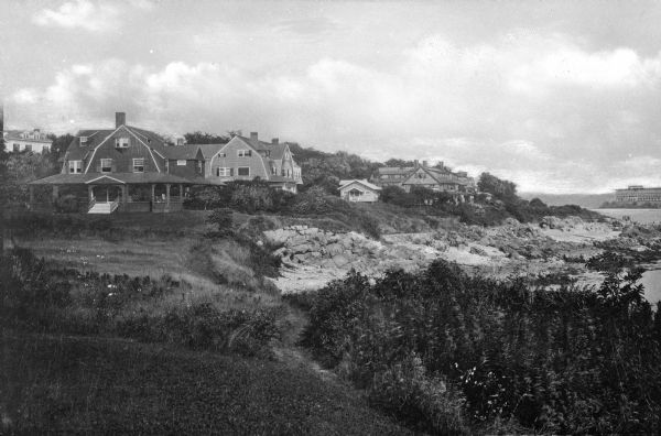 View along top of cliff toward cottages along a rocky coast. A large building is in the distance on the right.