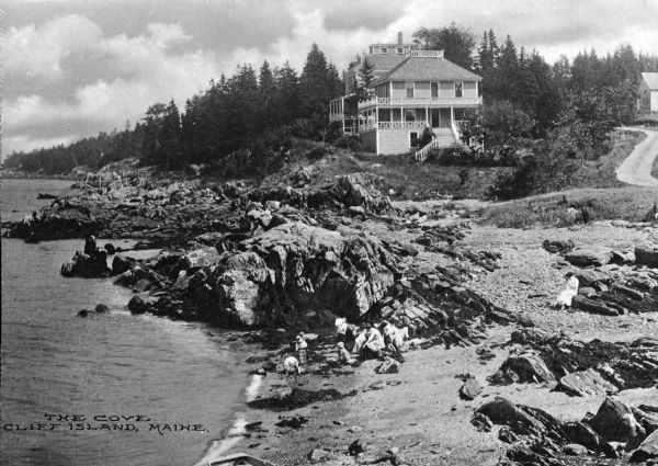 Elevated view of rocky shoreline, featuring a large building on a hill in the background. People are along the shoreline in the foreground. The caption reads: "The Cove, Cliff Island, Maine."