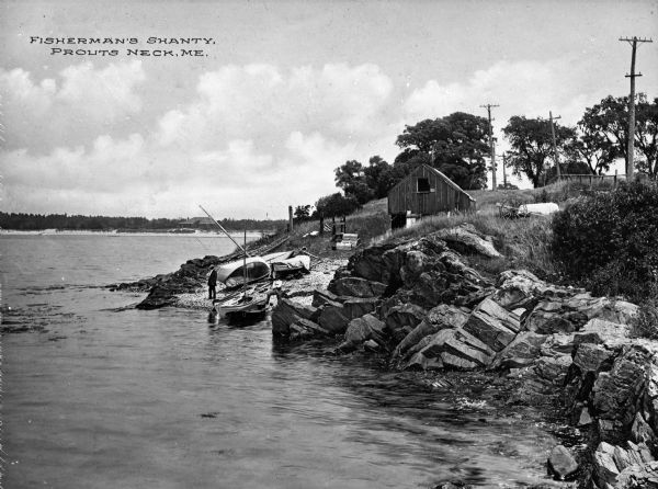 Elevated view of a fisherman's shanty along the shoreline surrounded by rocks. At the shoreline is a man and numerous boats pulled ashore. Caption reads: "Fisherman's Shanty. Prouts Neck. ME."