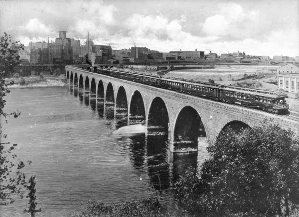Elevated view of an Oriental Limited train crossing The Stone Arch Bridge over a river. The bridge was built by The Great Northern Railway in 1883. There is a view of the central business district of the city in the distance.