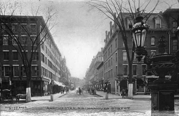 View up Hanover Street, featuring storefronts, horse-drawn carriages, a lamppost, and pedestrians. Caption reads: "Manchester, N.H., Hanover Street."