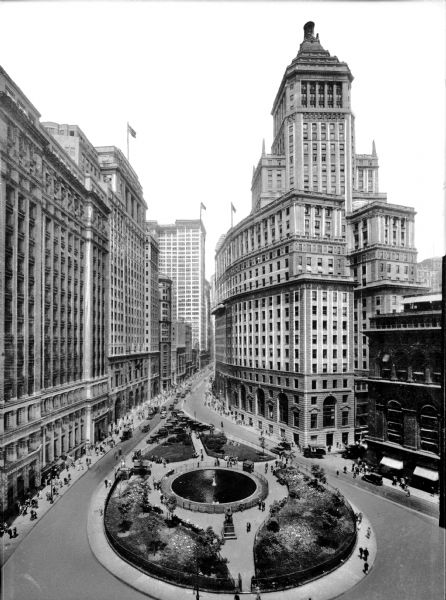Elevated view of a street scene in downtown, featuring a small park with a fountain, and many pedestrians among tall buildings.