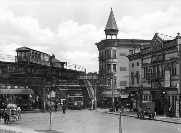 A view of Gotham Theater and a surrounding street scene, featuring trolleys, horse-drawn vehicles, pedestrians, and an elevated railway.