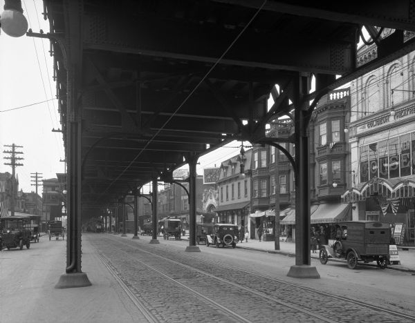 A view beneath an elevated railway at a market on 60th Street featuring pedestrians, storefronts, and cars.