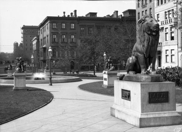 View of the park at Mt. Vernon Place, with a lion statue honoring the French artist Antoine-Louis Barye in the foreground, and a fountain and four additional statues in the rest of the park. In the background are buildings.