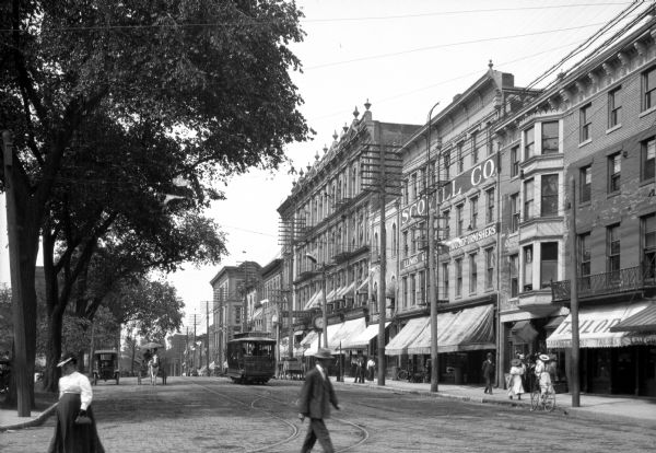 View of the 5th Street business district, featuring pedestrians, a trolley, an automobile, storefronts, and a horse-drawn carriage.