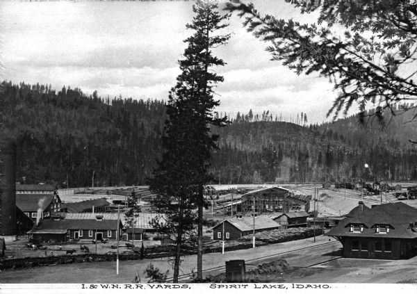 Elevated view of the railroad shipping yards of the Idaho and Washington Northern Railway. Included in the image are numerous buildings, workers, trees, and a background consisting of the surrounding tree-lined ridge. Caption reads: "I & W.N. R.R. Yards, Spirit Lake, Idaho."