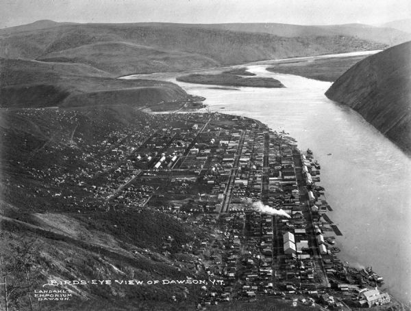 Aerial view of Dawson, Yukon Territory, Canada, from a surrounding hilltop, including a view of the distant hilltops and the adjacent Yukon river. Caption reads: "Bird's-Eye View of Dawson, Y.T."
