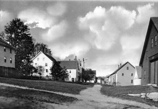 A low-level perspective looking down a sidewalk in the Shaker community, which was organized in 1794. There are numerous houses on both sides, and a windmill is in the distance.