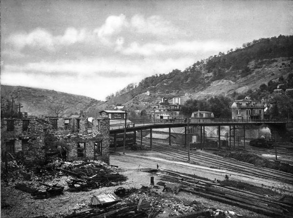 Elevated view of the Washington Bridge, crossing over numerous railroad tracks. In the foreground is a train engine, dwellings, and a dilapidated brick structure. In the background are hills.