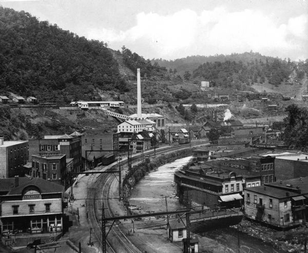 Elevated view of the city, which includes a factory, numerous buildings, railroad tracks, a bridge, Elizabeth river, and hills in the background.