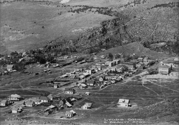 Elevated view of the small town in a rocky and hilly area including houses, roads and the surrounding empty landscape. Caption reads: "Lyons, Colo. A Beauty Spot."