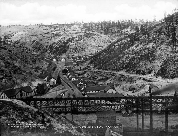 Elevated view of homes in a valley. In the foreground is a mining-car bridge, numerous buildings, and factory smokestacks. Caption reads: "Cambria, WYO."