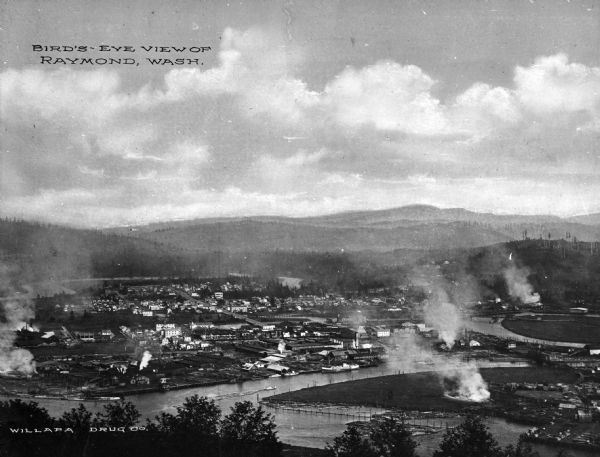 Distant elevated view of a lumbering town including a river and surrounding forest. Caption reads: "Bird's-Eye View of Raymond, Wash."