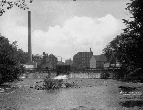 The Franklin Yextile Mills and T Steven's Company seen from the Winnehesauke River. View includes the factory dam, a smokestack and tall factory buildings.
