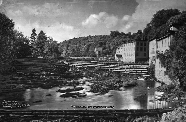 Elevated view of the Black River at low water, showing exposed stocks and wing dams with waterfront buildings and forested hills in the background. Caption reads: "River at Low Water."