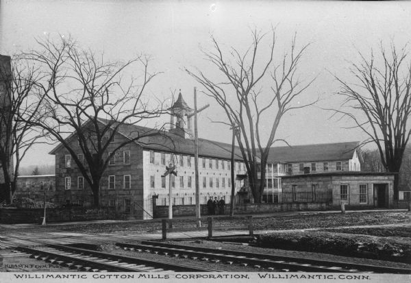 View of Willimantic cotton mills across railroad tracks with three men standing in front. Caption reads: "Willimantic Cotton Mills Corporation, Willimantic, Conn."