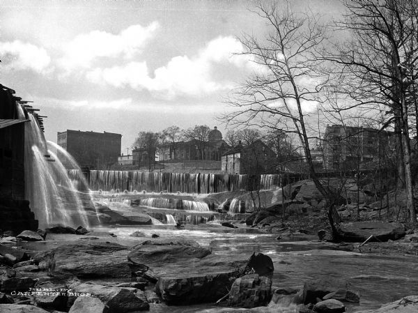 View of a mill dam and falls on the Saluda River with buildings in the background.