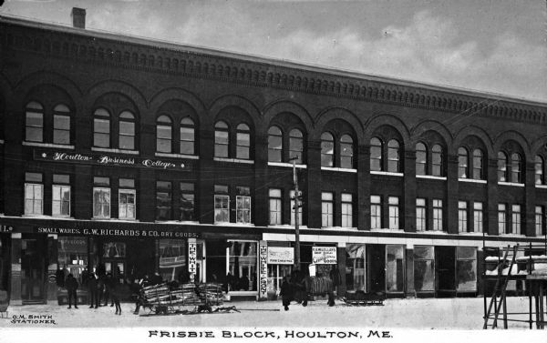 Frisbie Block. Row of small shops and businesses in the same large brick building, with a horse-drawn sleigh of firewood in front. Caption reads: "Frisbie Block, Houlton, ME."