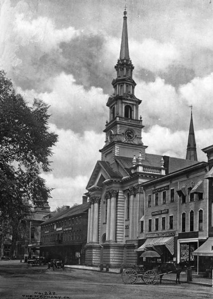 View of the United Church of Christ from across the street, including nearby storefronts as well as horse-drawn carriages and cars.