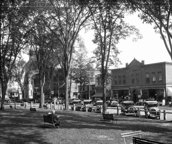 View of main street from a city park, including parked cars and store fronts.
