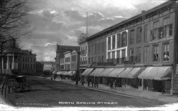 Row of shopfronts on North Spring Street in brick buildings with the Sussex county courthouse in the background. Caption reads: "North Spring Street."