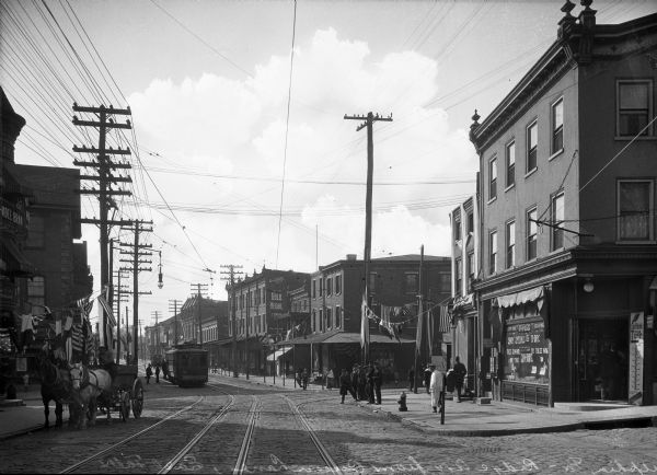 Street scene of Ridge Avenue from Queen Lake, East Falls. Features American flag decorations, people in the street, horse and carriages along the sides, as well as a cable car running down the middle of the street.