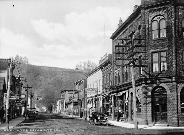 View down Main Street. Features storefronts and people of the town on the sidewalks. A few cars are parked at either side of the street. In the background is a large hill.