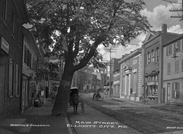 View down the cobblestone Main Street lined with storefronts. Features a large tree in the foreground as well as a horse-drawn carriage parked next to a restaurant. Caption reads: "Main Street, Ellicott City, MD."