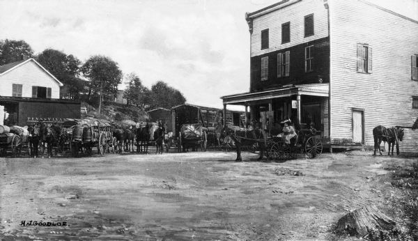 People with horses and wagons are congregated at the railroad depot.