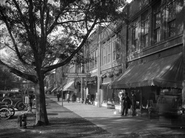 Street scene featuring Frierson's Pharmacy in the center. People walk the sidewalks and cars are parked in front of the row of stores.