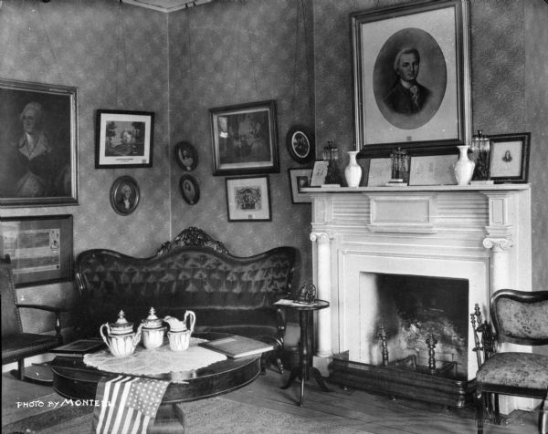 Interior of "Meadow Garden," home of Governor George Wallon. The living room is decorated with portraits, paintings, and other ornaments. Tea is set at the table.