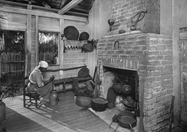 A woman is sitting by a fireplace churning butter, and seen through a window behind her is a man standing by a fence outdoors. Surrounding the fireplace are metal pots and pans, and baskets and carved gourds are displayed on shelves.