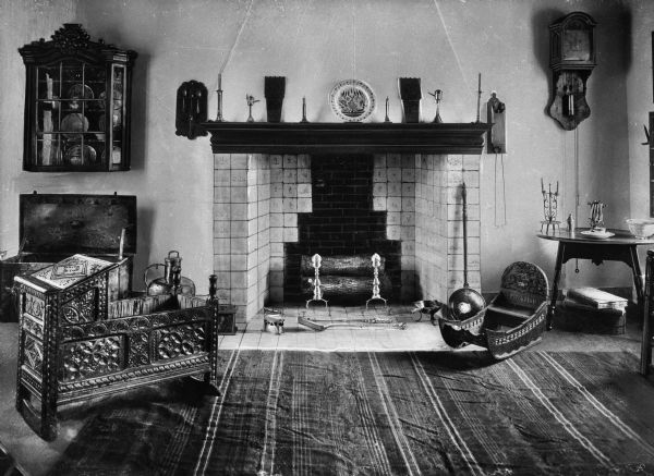 Interior view of the Dutch room of the Van Cortlandt house, featuring a fireplace, two ornately carved cradles, a wall clock, table, an open chest, polished metal implements and a display case of dishes.