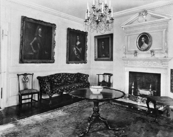 Interior view of a parlor in the Van Cortlandt House. Four portraits line the walls and a chandelier hangs above a table with a bowl on it in the center of the room. A fireplace is on the right.