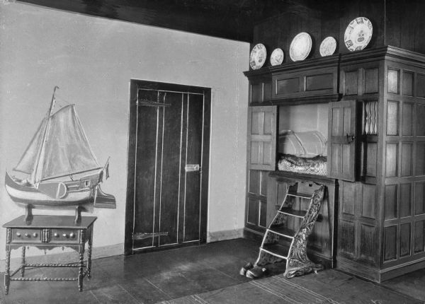 Interior view of a Dutch style bedroom at the Van Cortlandt House which features an enclosed paneled bed compartment with a set of wooden steps set in front. In the center is a door, and on the left is a model sailboat on a table. Ceramic plates are displayed along the top of the bed compartment.