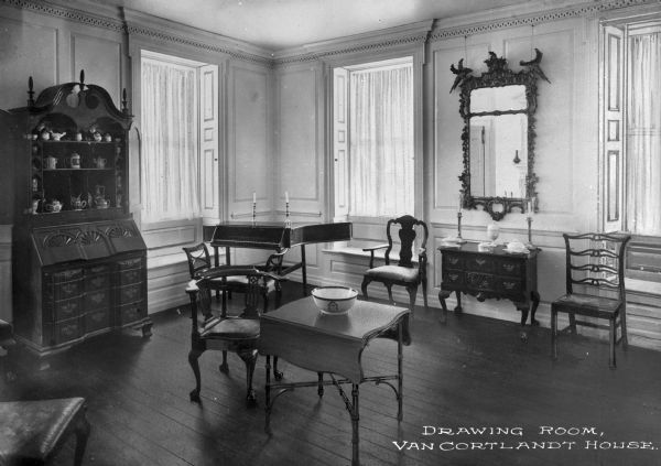 Interior view of the drawing room at the Van Cortlandt house which features bay windows with built in benches, ornately carved furniture, a harpsichord in the corner, and a large mirror in an elaborate frame. Caption reads: "Drawing Room, Van Cortlandt House."