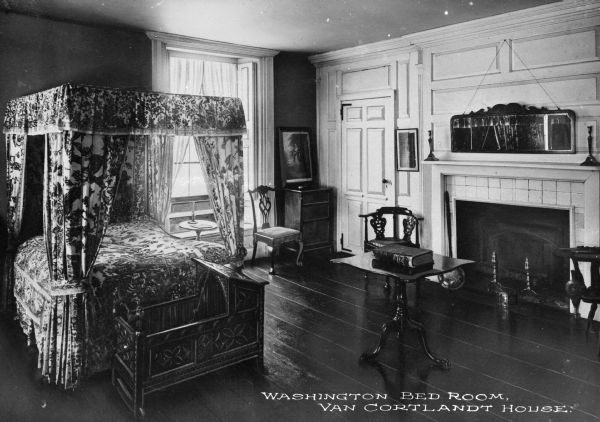 Interior view of the Washington bedroom at the Van Cortlandt house featuring a fireplace, canopied bed, with a Dutch-style carved cradle at the foot, other carved furniture, and hanging artwork. Caption reads: "Washington Bed Room, Van Cortlandt House."