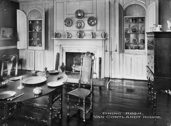 Interior view of the Van Cortlandt house dining room featuring a table set with shining metal tableware, decorative plates on the wall, and open cabinets exposing the china and tea sets within. Caption reads: "Dining Room, Van Cortlandt House."
