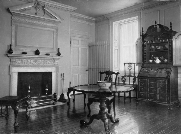 Interior view of the Van Cortlandt house drawing room with hardwood floor. Features a fireplace with a gleaming metal spark-guard, an ornately carved table, chairs, and desk. Ceramics including bowls, teapots and vases are displayed throughout the room.
