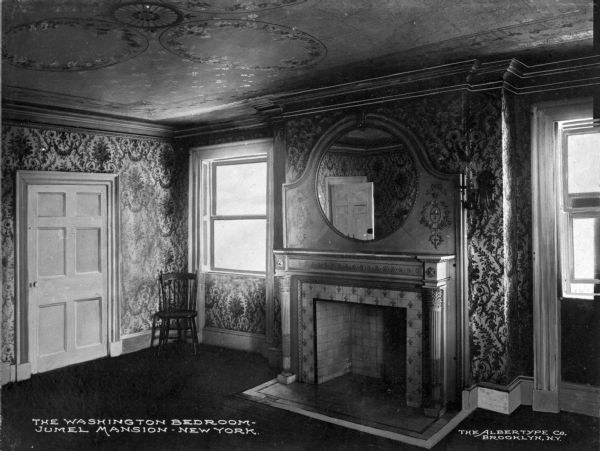 Interior view of the Morris-Jumel Mansion which served, at different times, as headquarters to both forces during the Revolutionary war.  This room features a chair in the corner, an ornate fireplace with a large, round mirror above, intricate wallpaper and decorative painting on the ceiling. Caption reads: The Washington Bedroom - Jumel Mansion, New York."