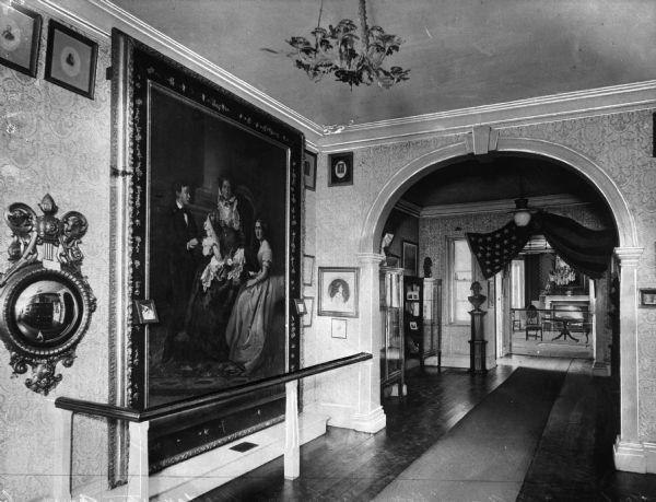 Interior view of the Morris-Jumel Mansion which served, at different times, as headquarters to both forces during the Revolutionary war.  This view of the entrance hall features hanging artwork, including a large portrait on the wall on the left behind a railing, chandeliers, sculptures, and an American flag above the open doorway at the back of the image.