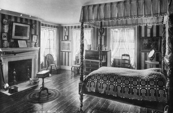 Interior view of the Morris-Jumel Mansion which served, at different times, as headquarters to both forces during the Revolutionary war.  This view of Washington's bedroom features a carved canopied bed, an open writing desk, a gun hanging above the fireplace, and artwork and other objects throughout the room.